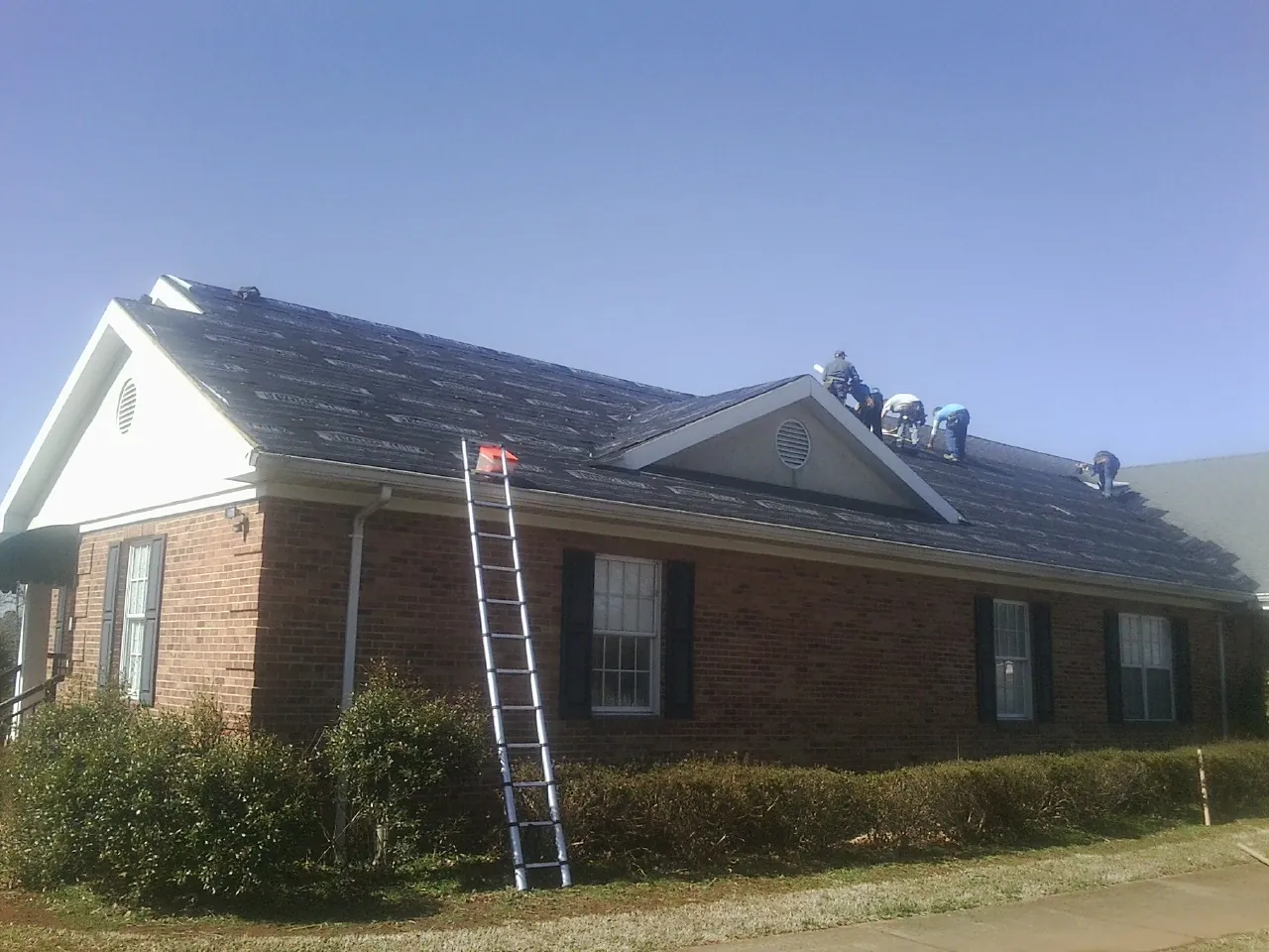 Crew tearing shingles off a brick ranch roof with a ladder set — Metal Roof Repair work by our crew in Holton