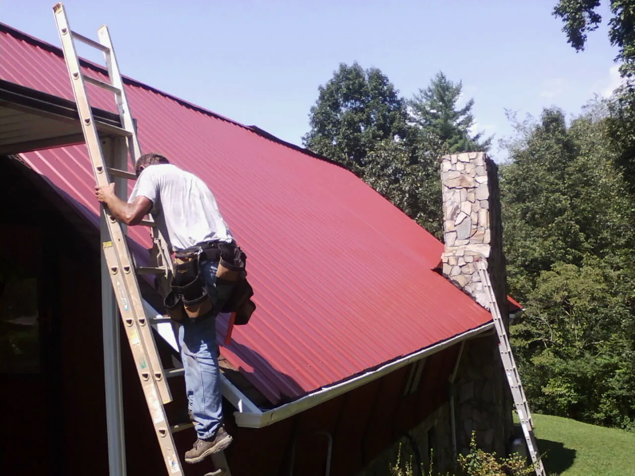 Roofer on a ladder beside a red metal roof and stone chimney — Soffit & Fascia Repair by Roofing Brothers in Holton